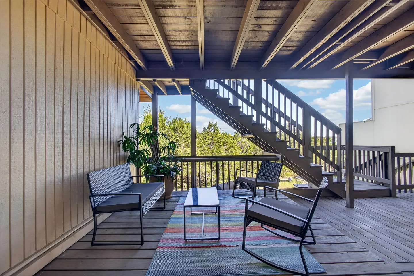 A covered wooden deck furnished with a bench, rocking chairs, a coffee table, and an area rug, with wooden stairs leading up.