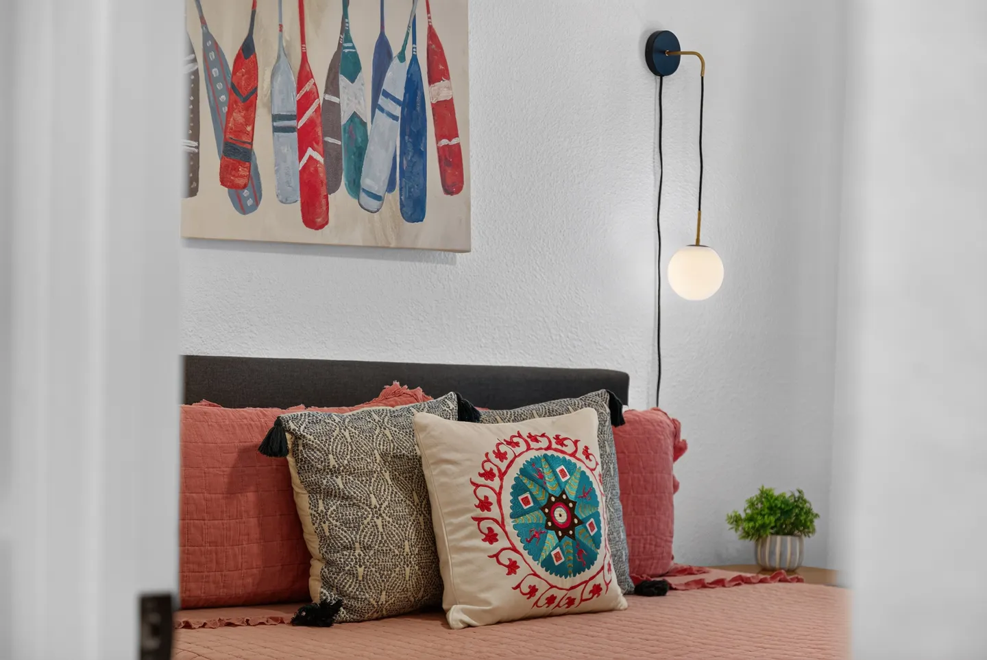 Close-up of a made bed with decorative patterned pillows, a wall sconce with a spherical bulb, and a wall painting depicting colorful boat paddles.