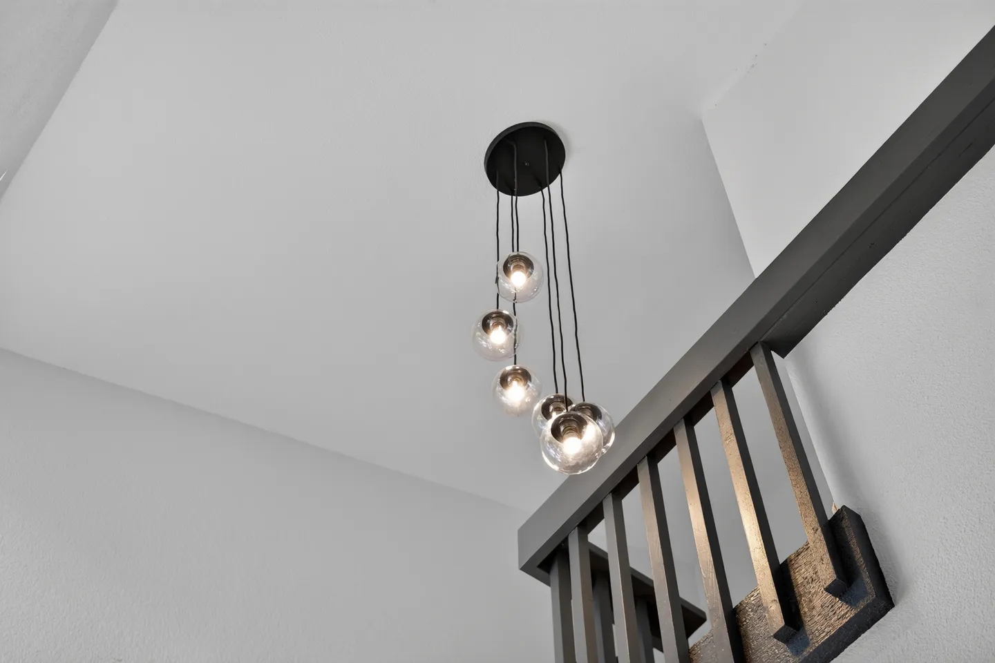 Low-angle view of a modern multi-bulb glass pendant light hanging near a dark wooden staircase railing against light gray walls.