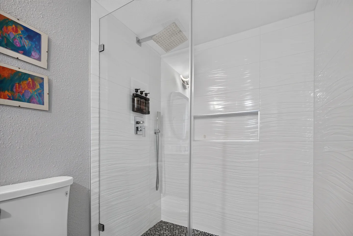 Walk-in shower with glass doors, textured white wall tile, rain shower head, handheld sprayer, and built-in niche next to a toilet.