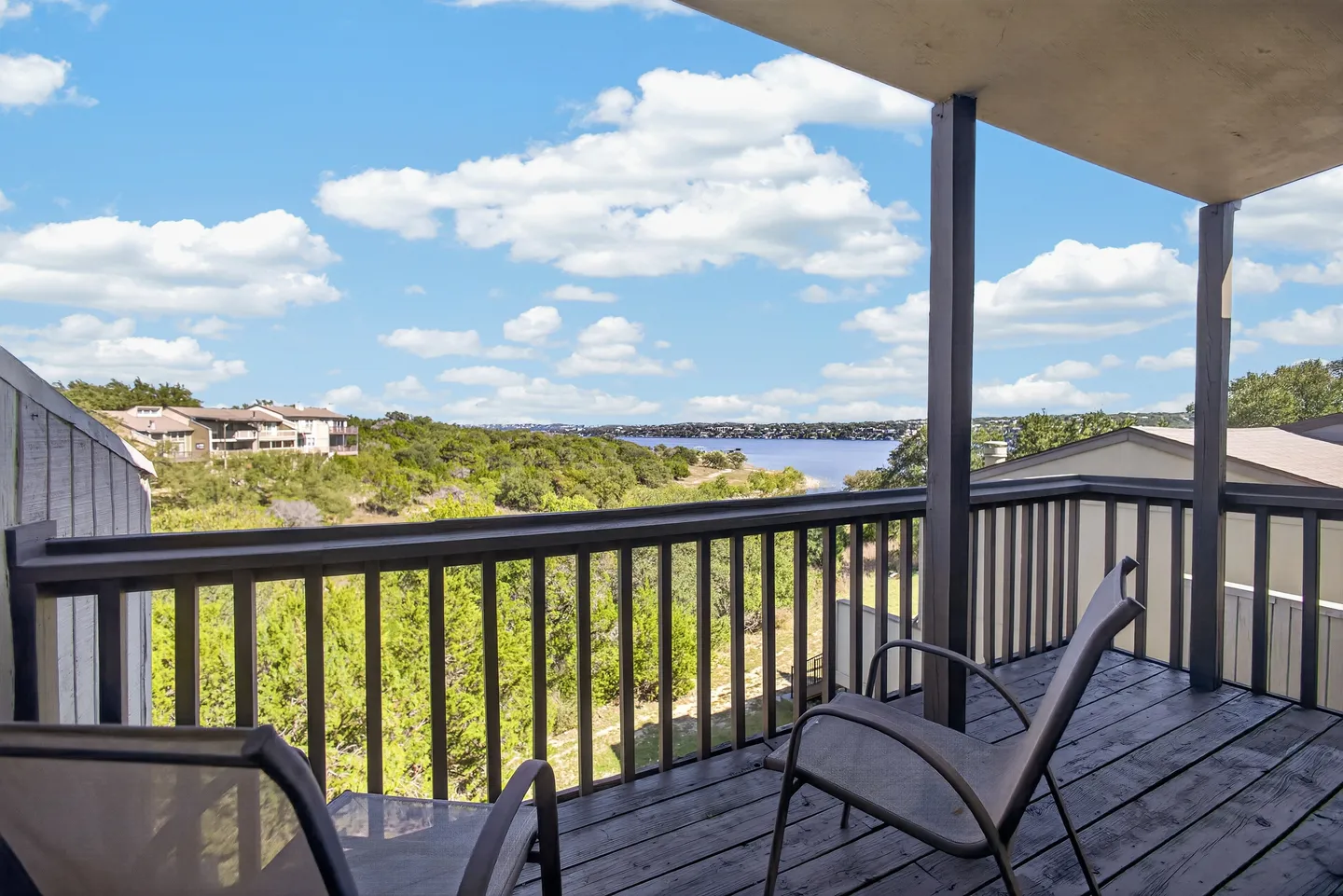 A covered wooden balcony with two chairs overlooking a body of water and rolling green hills under a partly cloudy sky.
