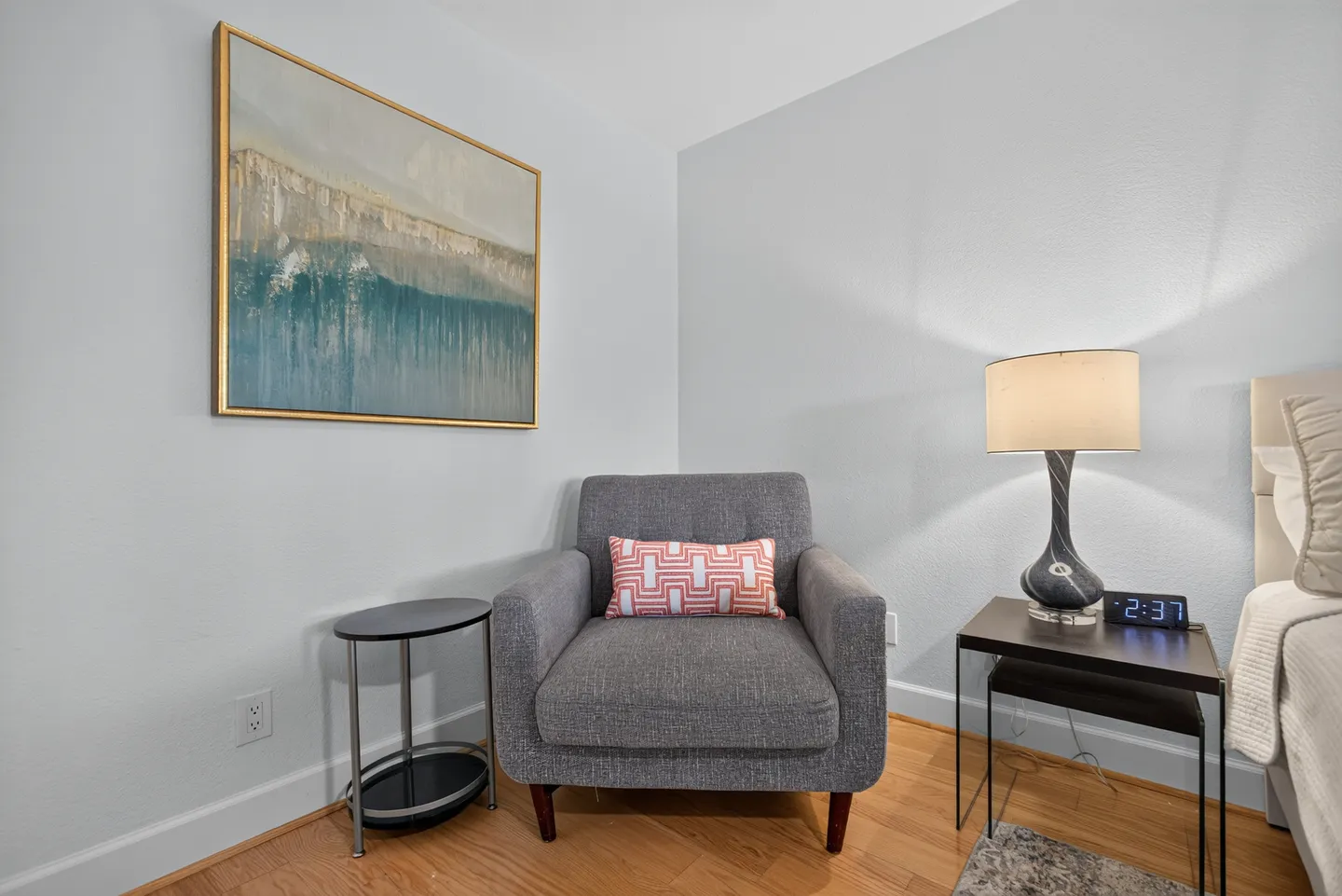 A corner sitting area in a bedroom featuring a gray armchair, two side tables, a framed painting, and an illuminated lamp.