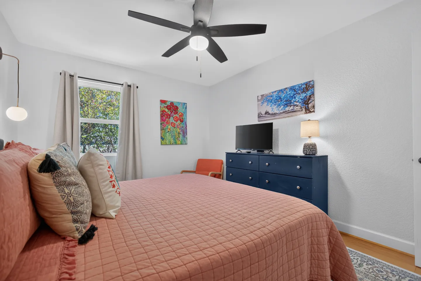 Bedroom with a bed made in a coral-pink quilt, a blue dresser with a television, and a window.