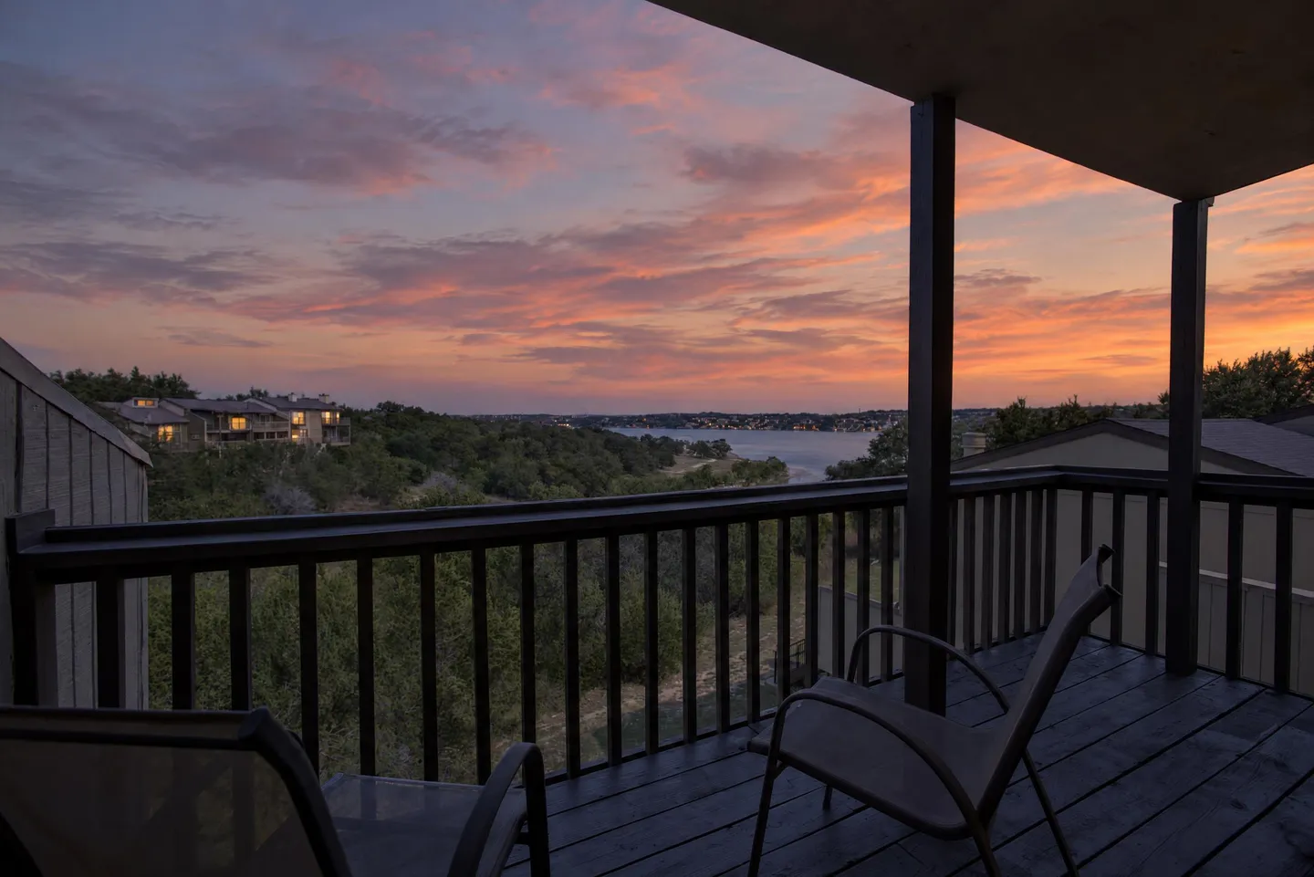 A covered deck with dark railings and chairs overlooks a sunset view of trees, houses, and a body of water in the distance.