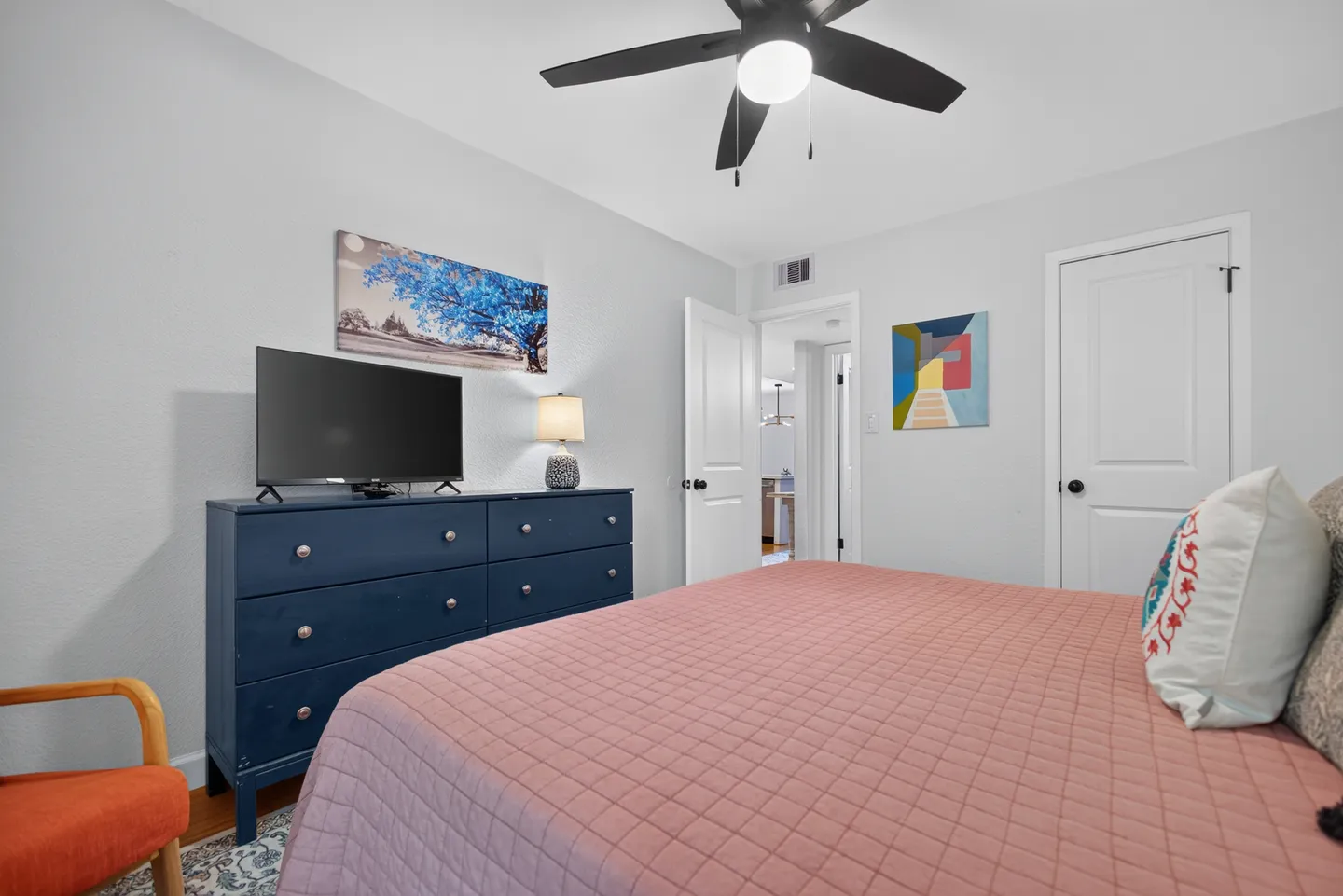 Bedroom featuring a bed with a pink quilted coverlet, a blue dresser with a mounted TV, and an open doorway leading to a hallway.