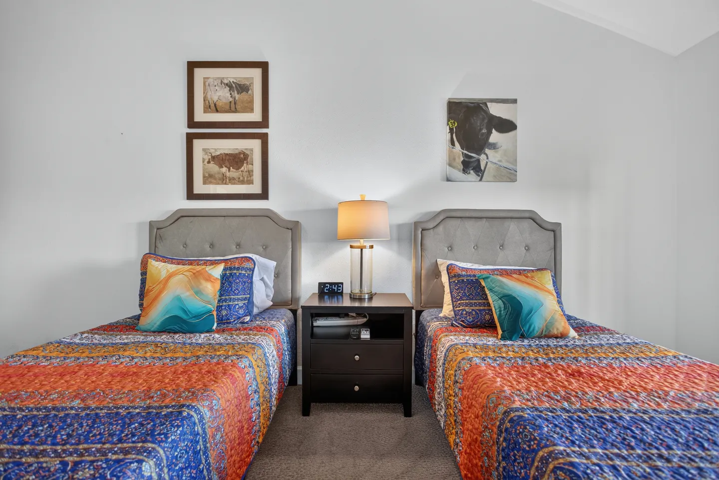 Bedroom with two twin beds featuring tufted gray headboards, vibrant multicolored quilts, and a shared nightstand with a lamp.