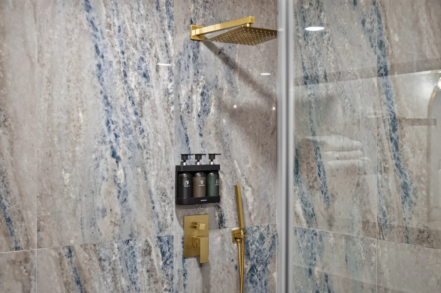 Close-up of a walk-in shower with blue and grey marbled wall tiles, gold fixtures, and wall-mounted dispenser bottles.