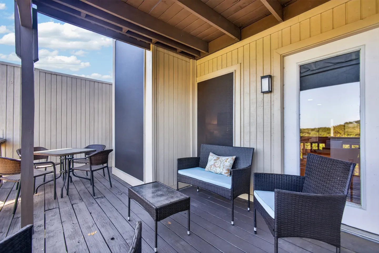 Covered outdoor deck with dark wicker seating, a small coffee table, and an outdoor dining table with chairs under an open sky.