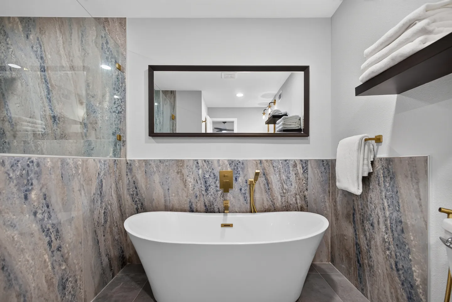 Freestanding white soaking tub against a blue-and-tan marble-look wall with gold fixtures and a framed mirror above.