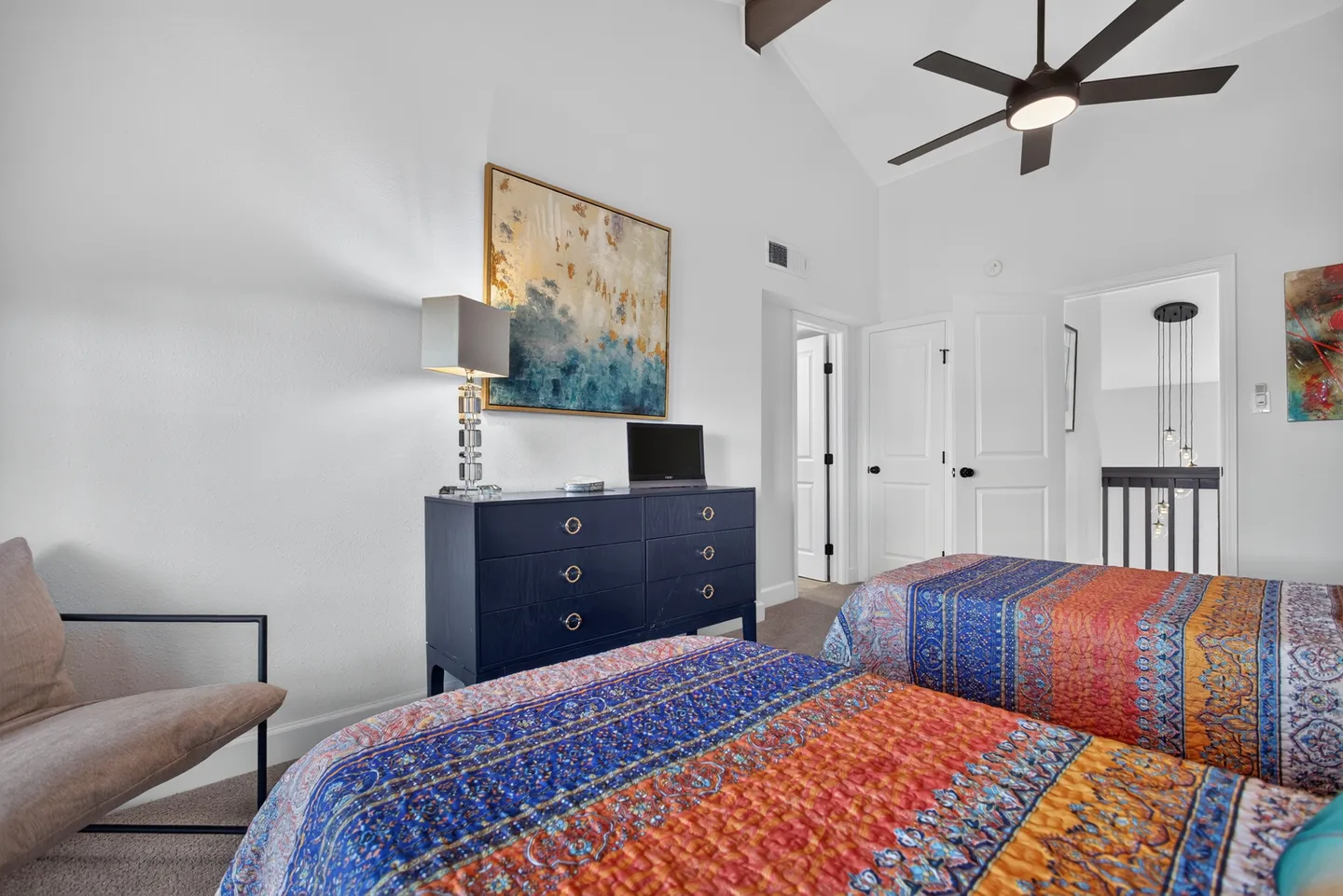 Bedroom with two twin beds dressed in colorful patterned quilts, a navy blue dresser, and a vaulted ceiling with a fan.