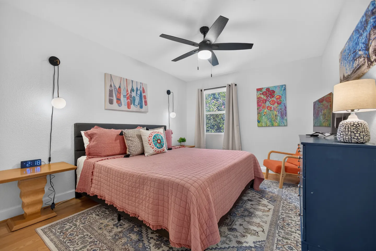 Bedroom with a bed made with a dusty pink quilt, flanked by nightstands, beneath a ceiling fan and across from a blue dresser with a TV.