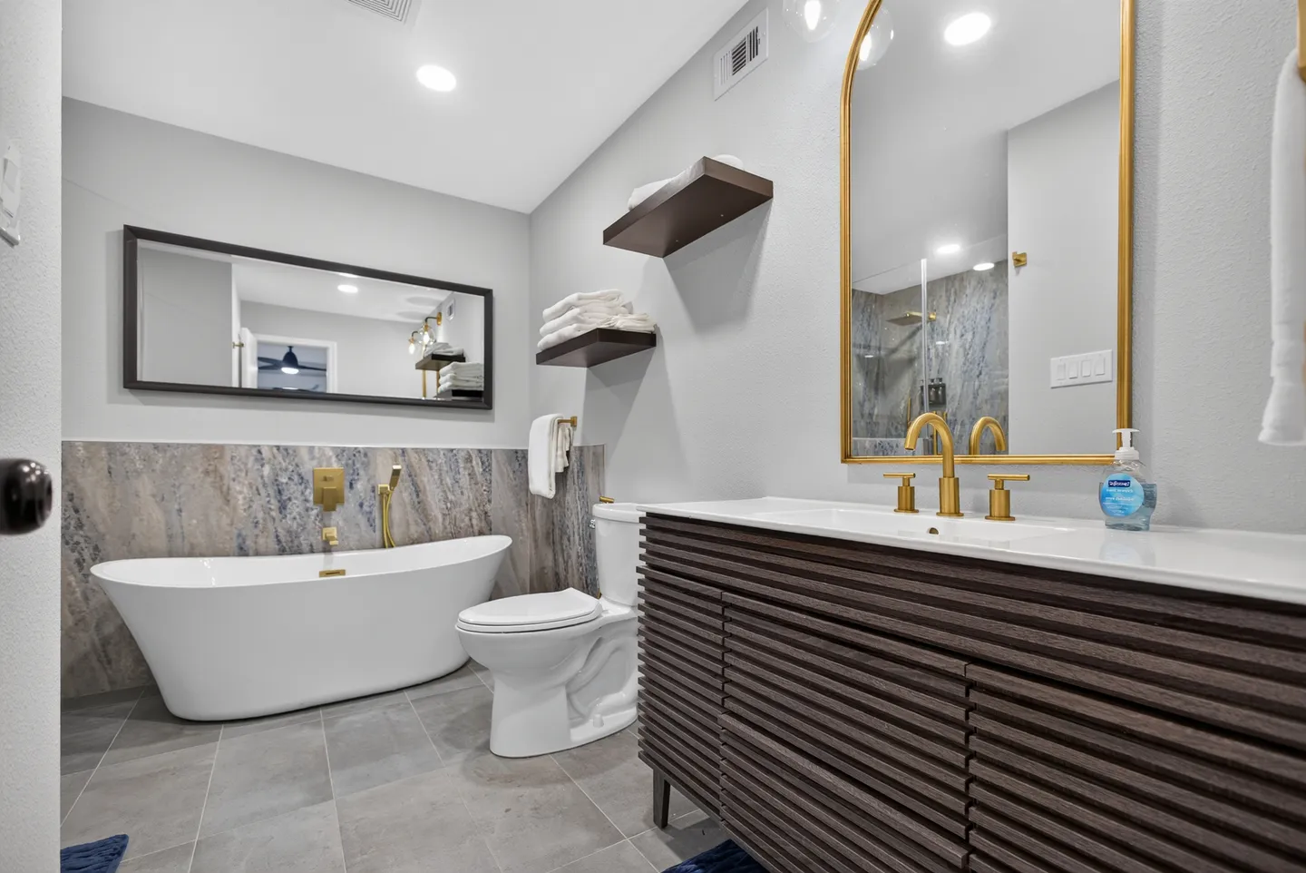 Bathroom featuring a freestanding white soaking tub, dark wood slatted vanity with gold faucet, arched gold mirror, and toilet.