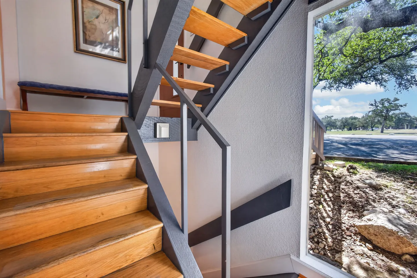 Open-tread wood staircase with dark gray metal railings next to a large window overlooking the outdoors.