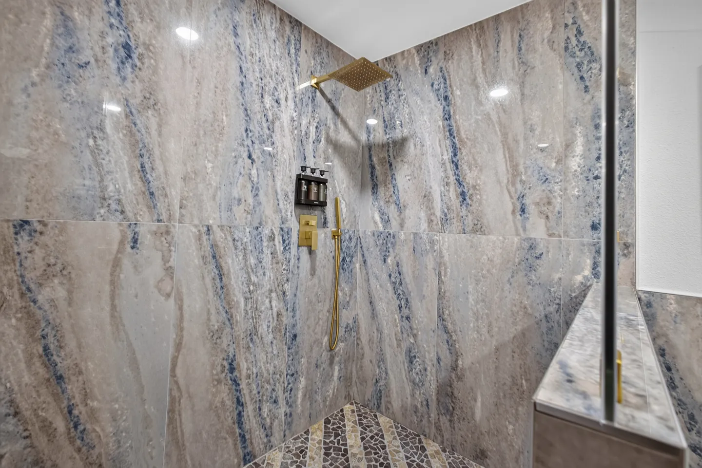 A walk-in shower with marble-look blue and gray wall tiles, gold-toned fixtures, and a pebble tile floor.
