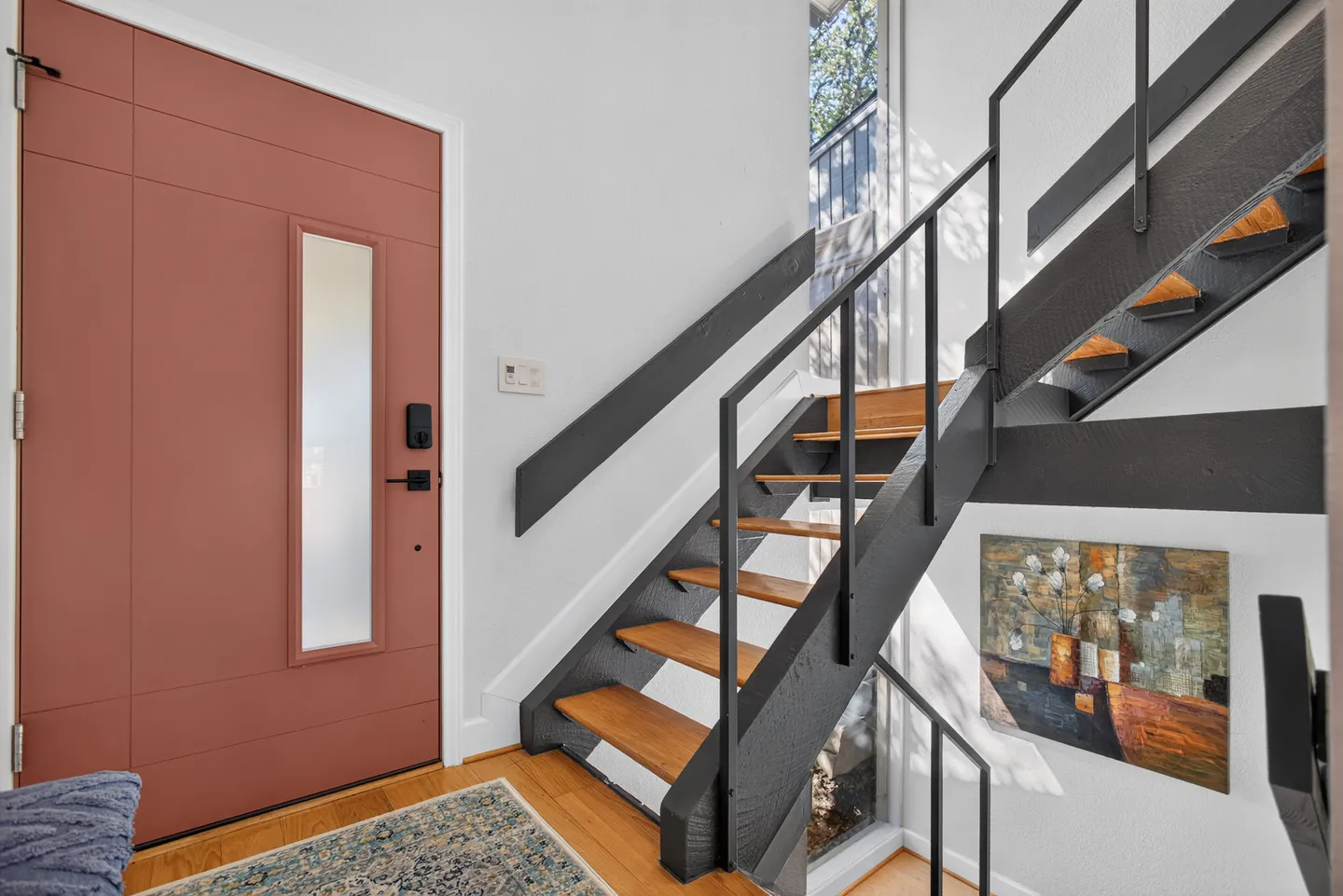 Entryway featuring a terracotta-colored front door with a frosted glass panel beside an open-tread staircase with black railings.