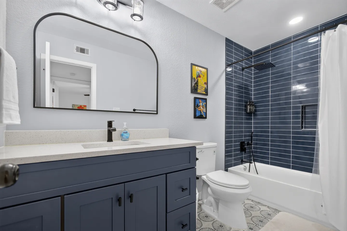 Bathroom with a dark blue vanity, an arched mirror, a toilet, patterned tile flooring, and a shower-tub combination featuring glossy blue subway tiles.