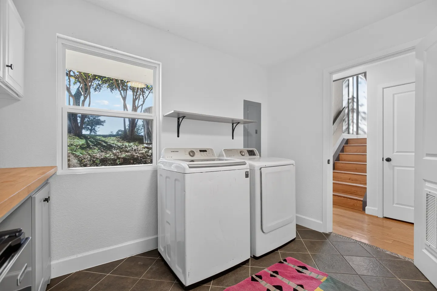 Laundry room with white washer and dryer, tiled floor, a window looking outside, and a view into a hallway with stairs.