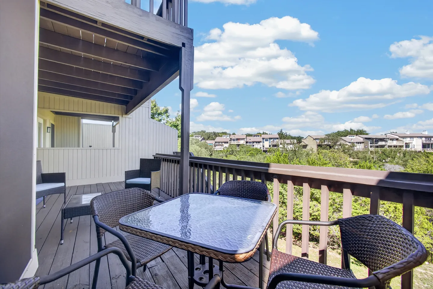 Outdoor wood deck with wicker armchairs, a glass-topped dining table, and views of surrounding buildings under a blue sky.