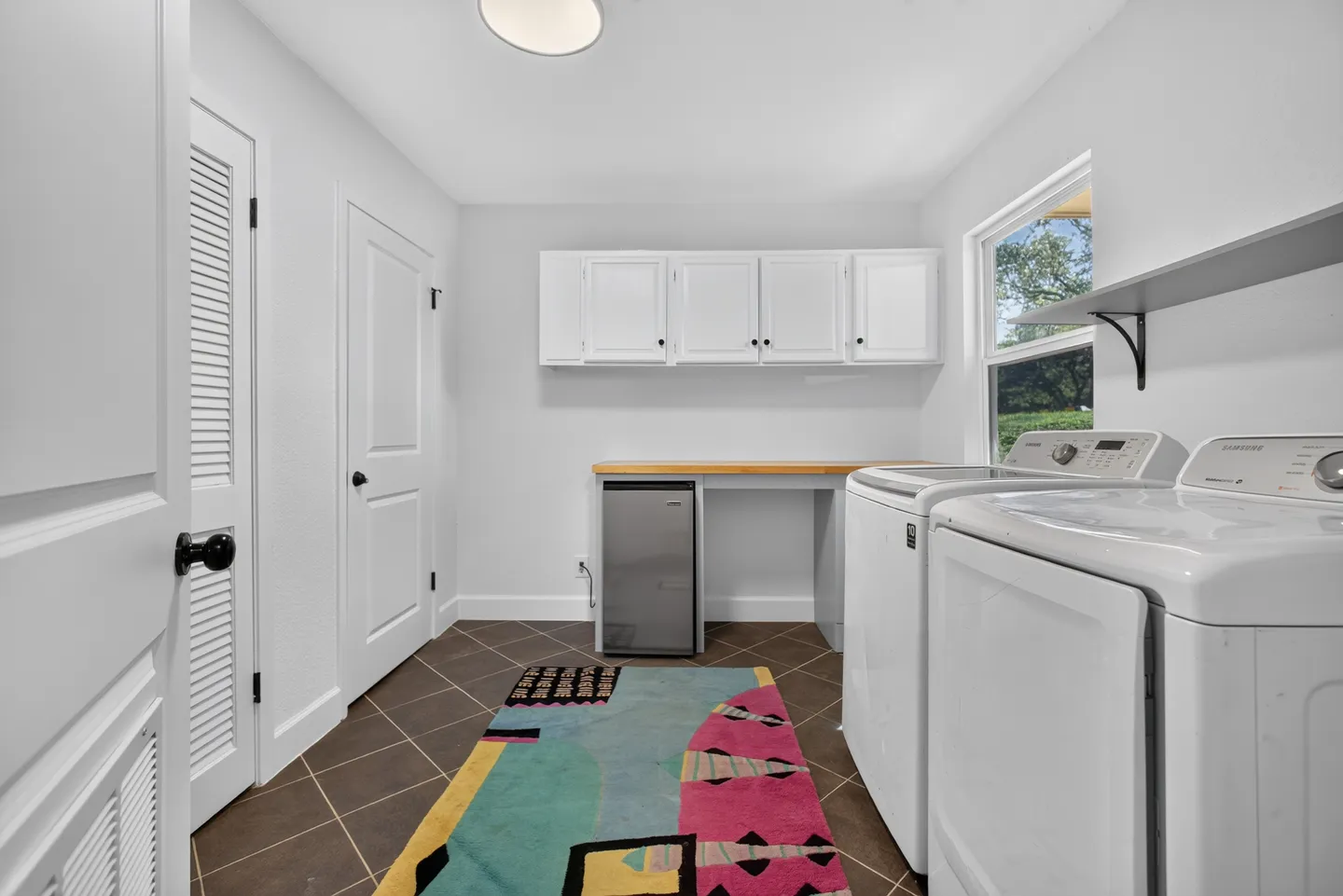 Laundry room with white walls, tiled floor, white washer and dryer, small beverage fridge under a wooden counter, and white upper cabinets.