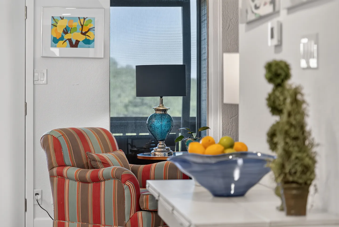 Indoor seating nook featuring a striped armchair, a blue glass lamp on a side table by a window, and citrus fruit in a bowl on a console.