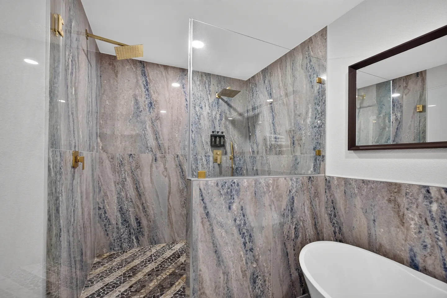 Bathroom showing a walk-in shower with patterned stone tile walls, gold fixtures, and a freestanding soaking tub.