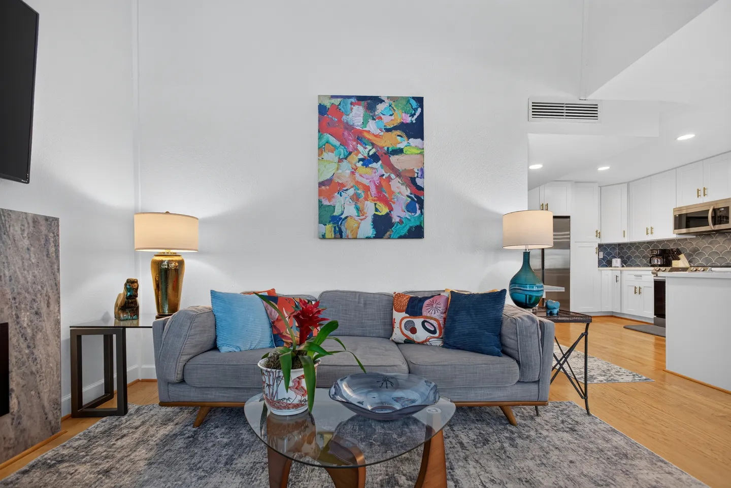 Living room featuring a grey sofa with colorful throw pillows, a glass coffee table, abstract art on the wall, and a view into the kitchen.