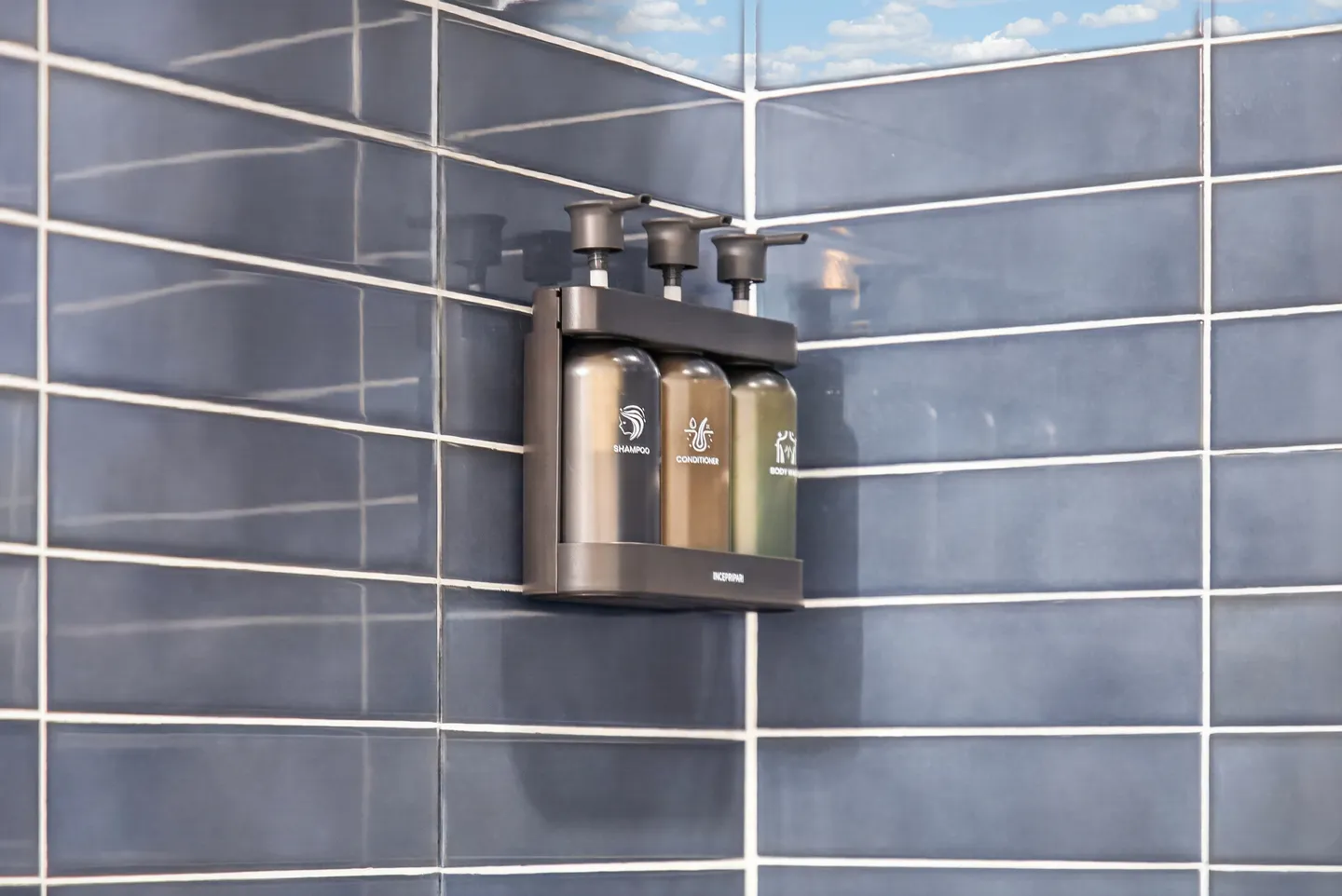 Close-up of a wall-mounted shampoo, conditioner, and body wash dispenser in a blue-tiled shower.