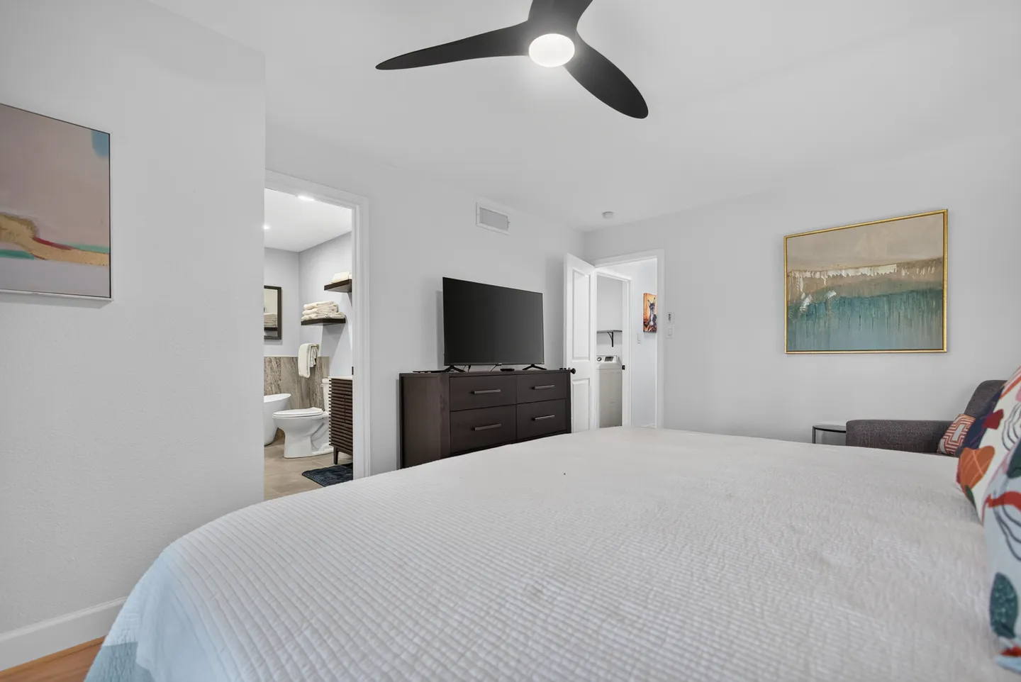 Bedroom with a white bed, ceiling fan, dark wood dresser, mounted television, and views into an ensuite bathroom and hallway.