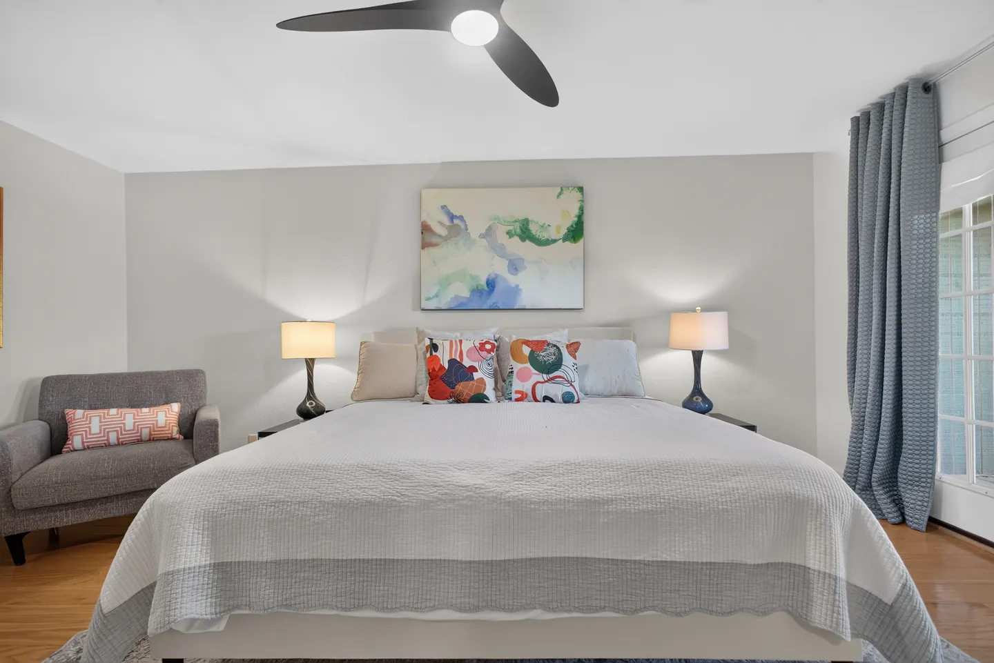 Bedroom with a large bed dressed in white and gray bedding, flanked by nightstands with lamps, a ceiling fan above, and an armchair to the side.