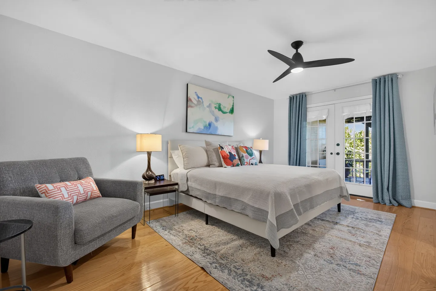 Bedroom with a made bed, patterned area rug over wood floors, an armchair, and French doors flanked by blue curtains.