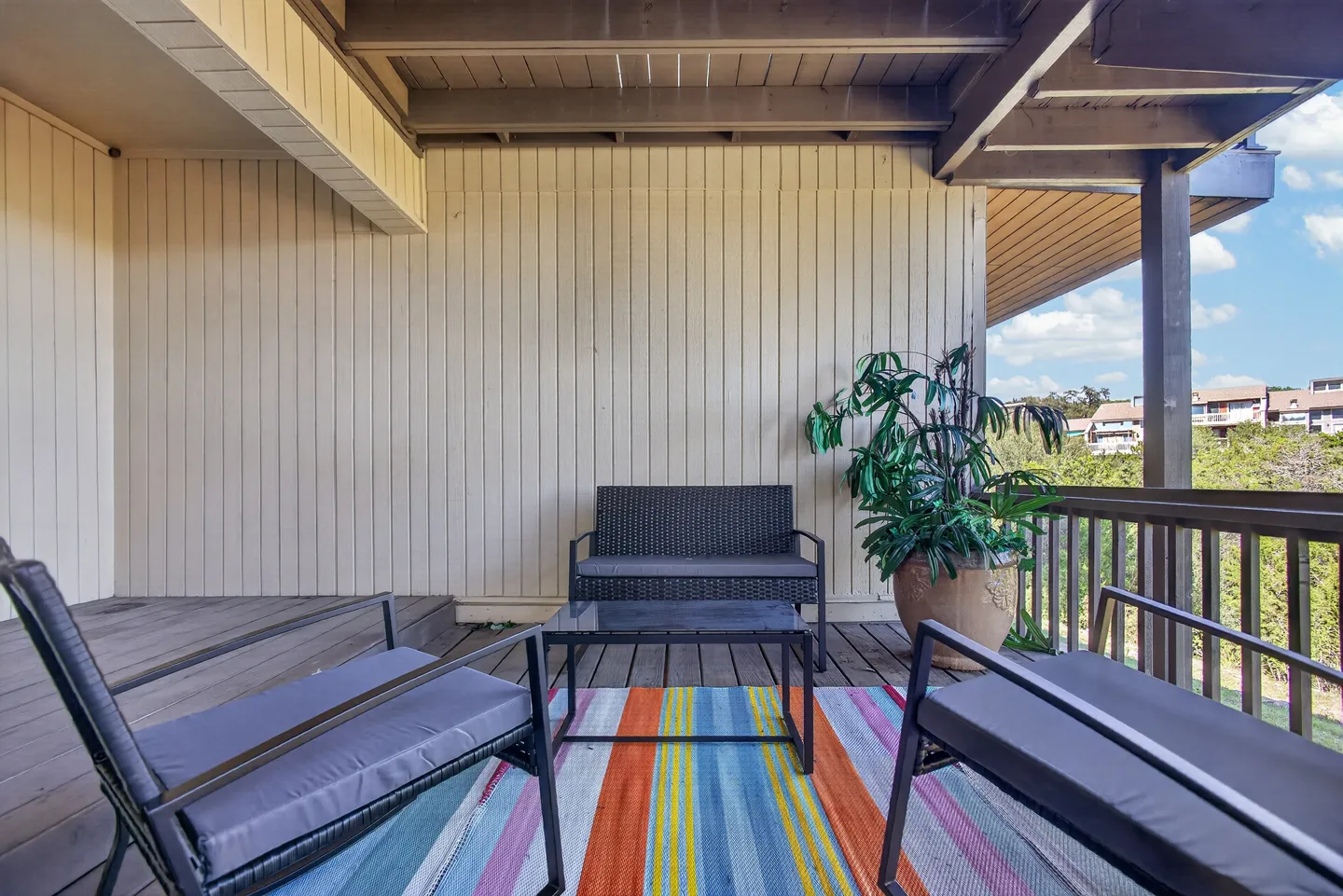 Covered deck with outdoor seating, a striped rug, a potted plant, and a railing overlooking trees and buildings under a blue sky.