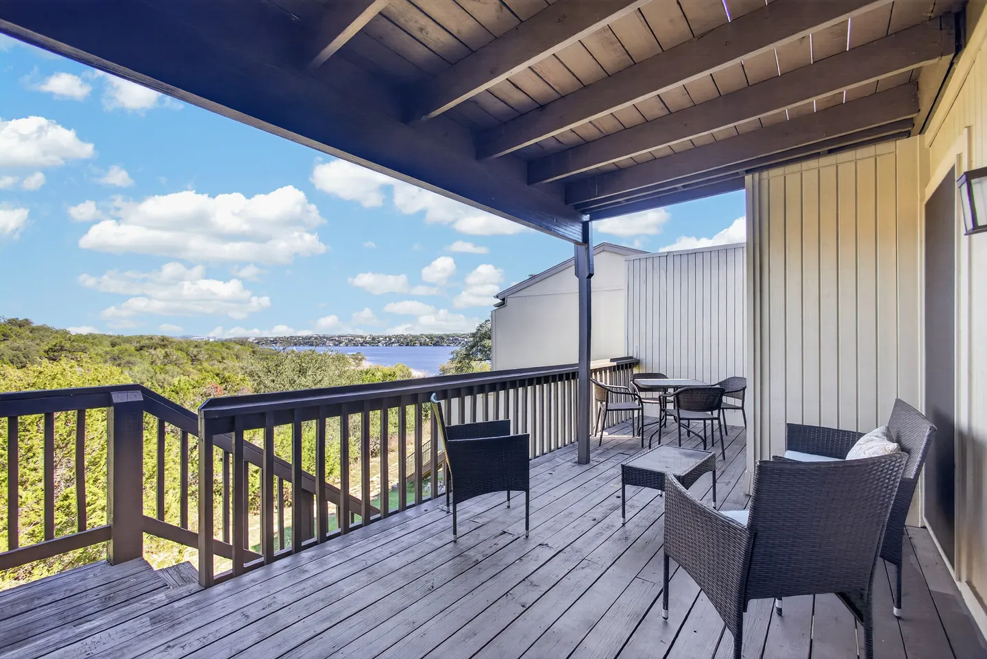 Covered wooden deck with outdoor seating overlooking green trees and a distant body of water under a blue sky.