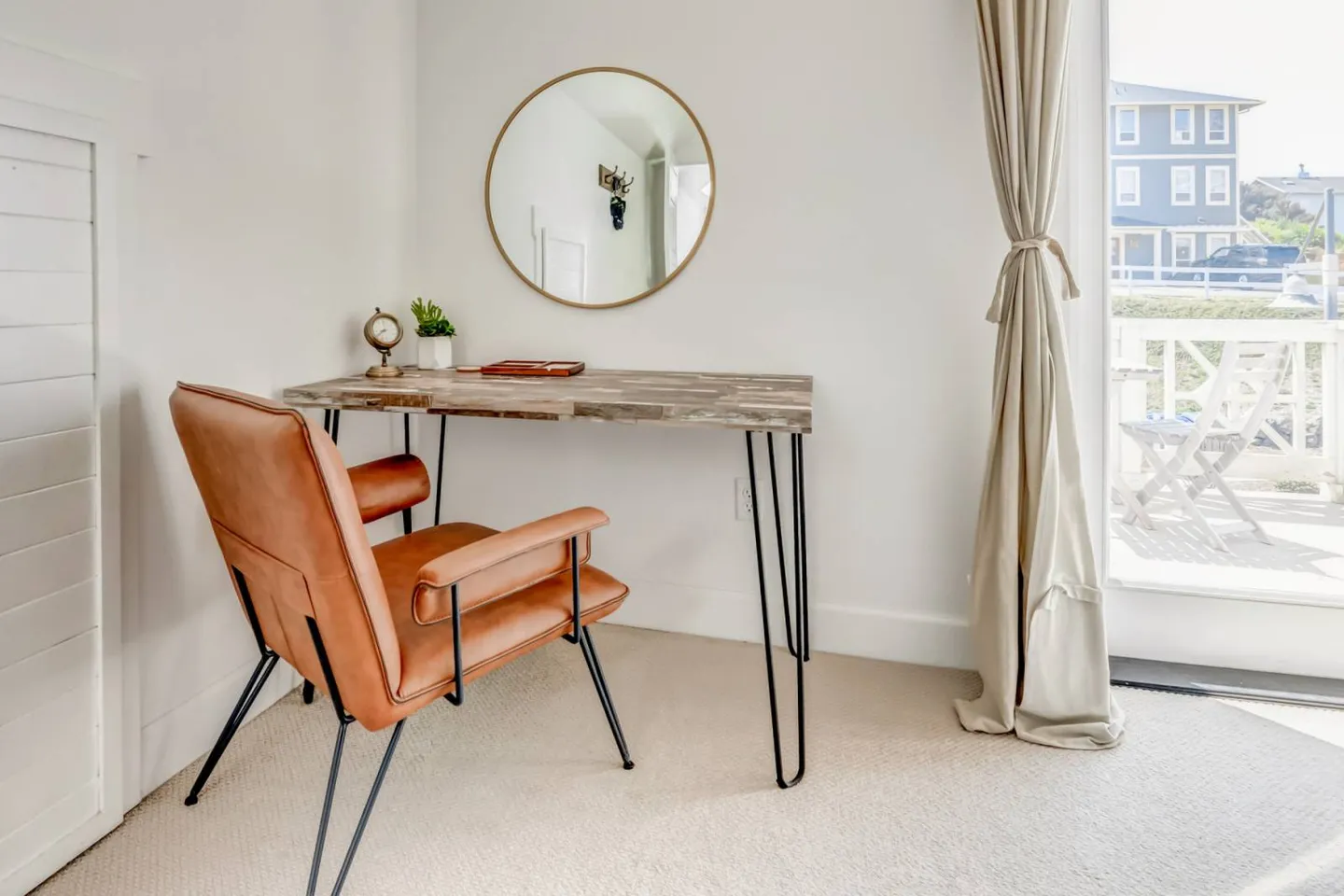 A dedicated workspace featuring a wood desk with hairpin legs, a brown leather armchair, and a circular mirror on a white wall next to a glass door.