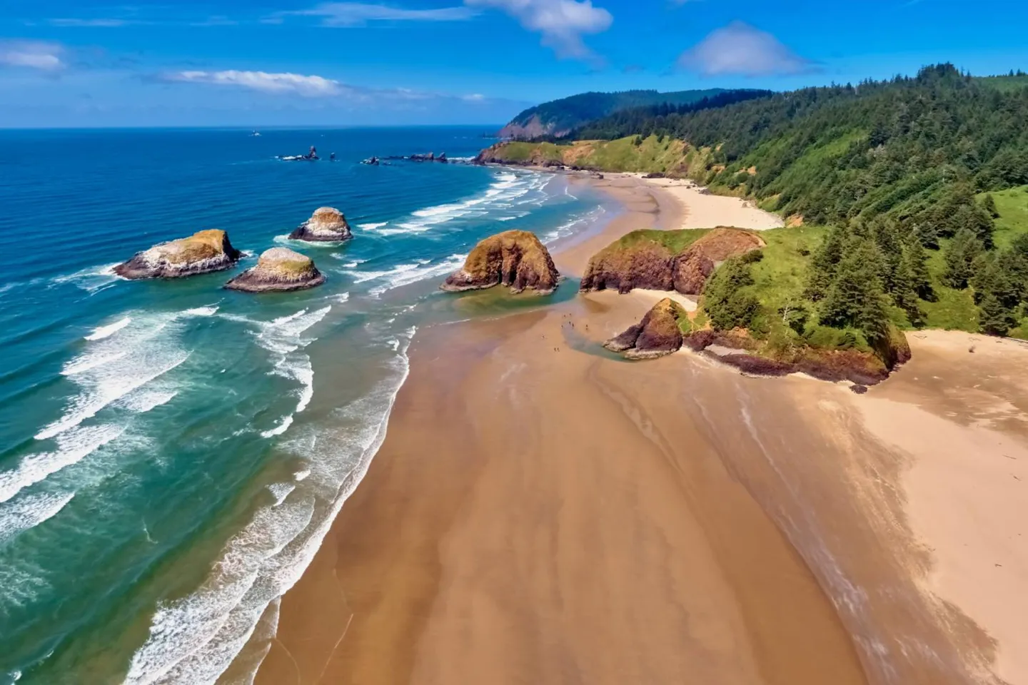 Aerial view of a wide sandy beach featuring large sea stack rock formations, rolling waves, and forested coastal cliffs under a blue sky.