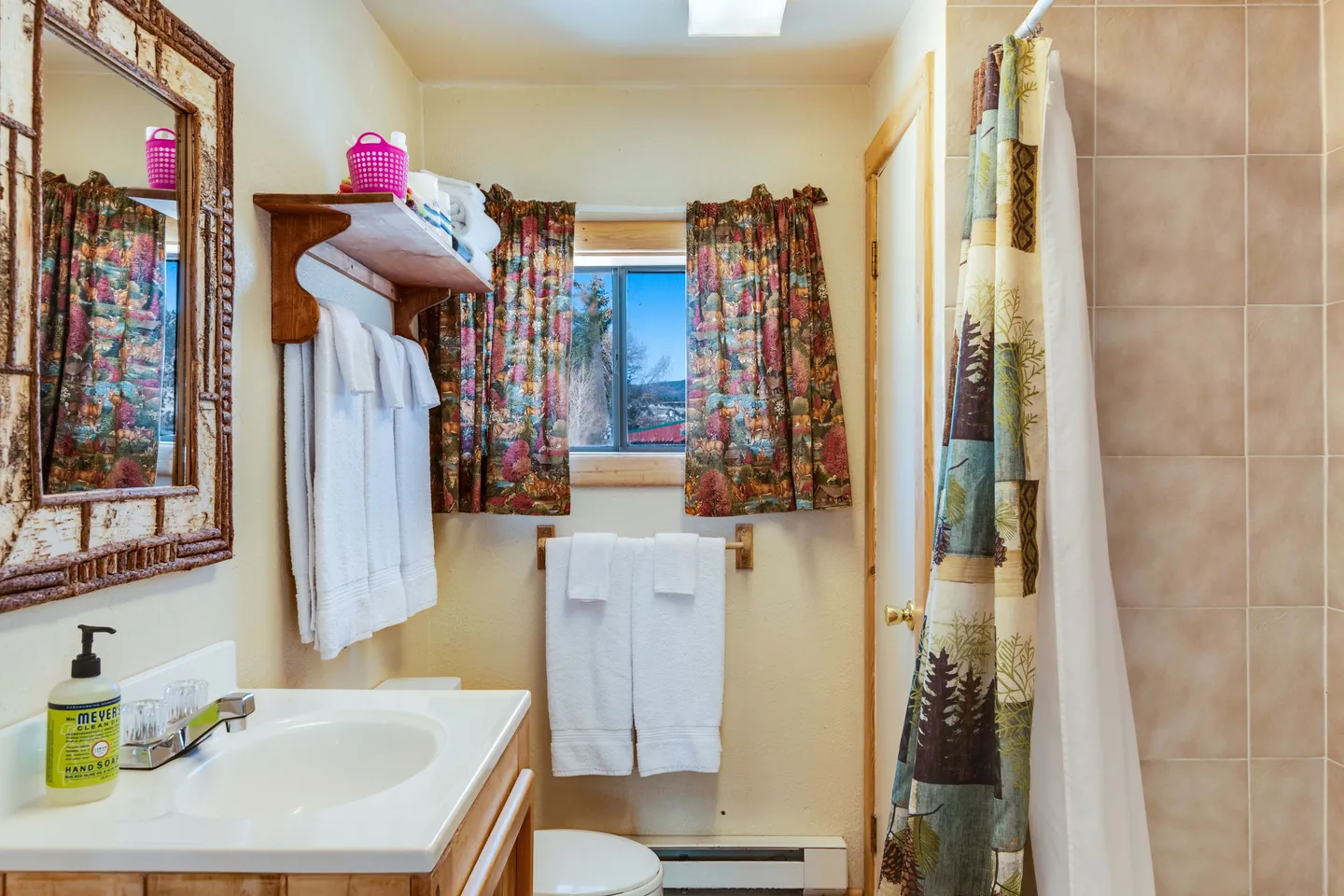 Bathroom with vanity, toilet, tiled shower with nature-themed curtain, and window with colorful curtains.