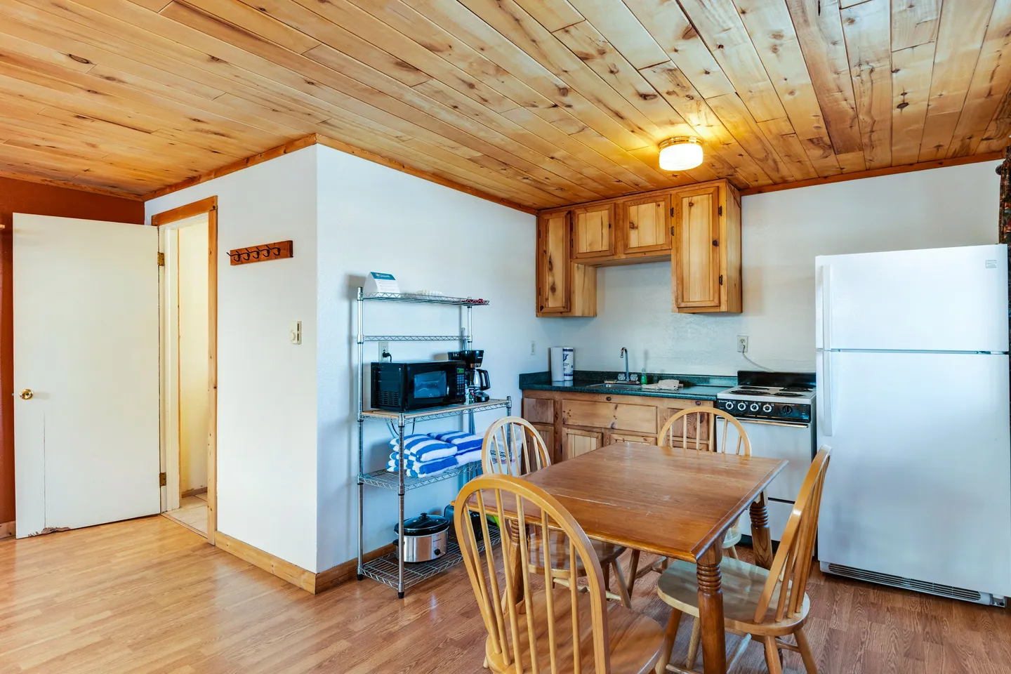 Kitchen and dining area with wood-paneled ceiling, wooden cabinets, dining table with chairs, full-size refrigerator, and wire shelving rack.