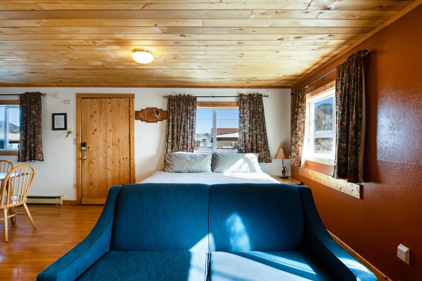 Studio interior showing a blue couch in the foreground, a bed by a window in the background, a wooden door, and a wood-paneled ceiling.