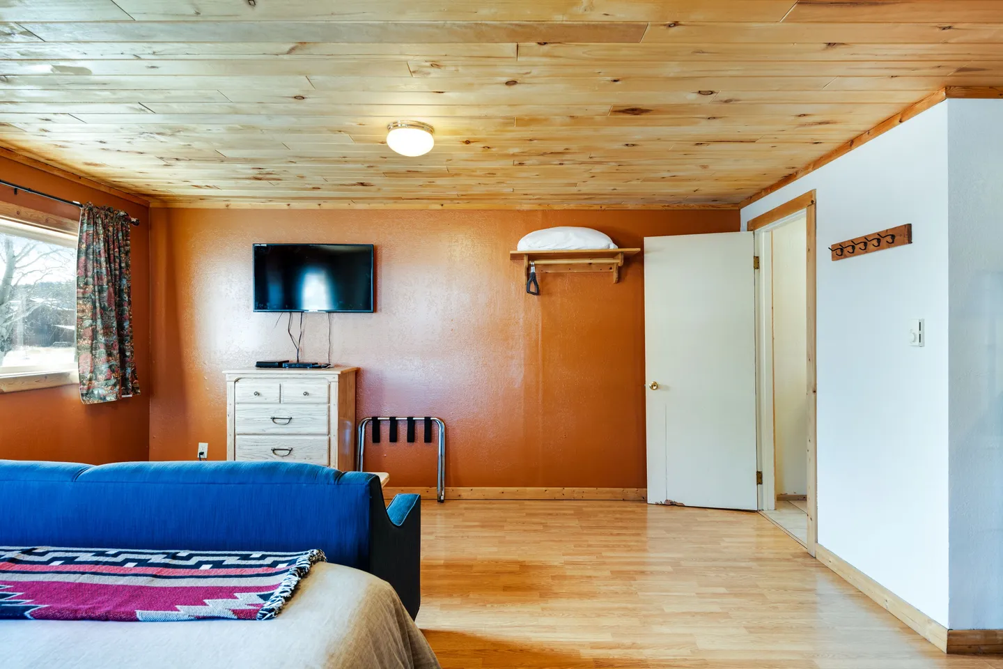 Bedroom featuring wood-paneled ceiling, light wood-look flooring, rust-orange accent wall with a wall-mounted TV over a small dresser, and an open doorway leading to another room.