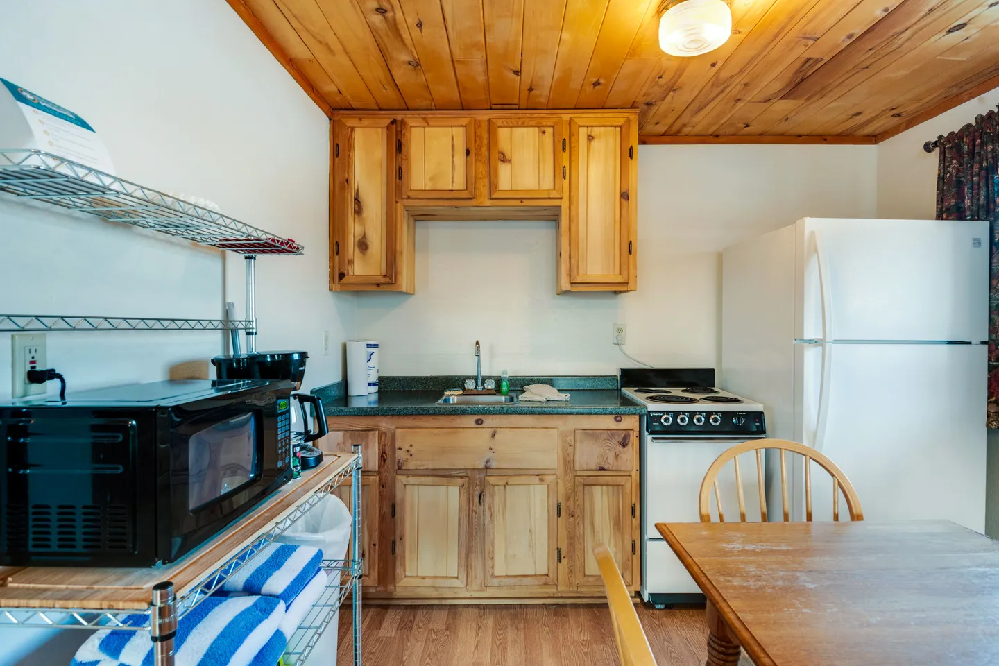 A compact kitchen featuring light knotty pine cabinets, a wood plank ceiling, an electric range, refrigerator, and an adjacent dining table.