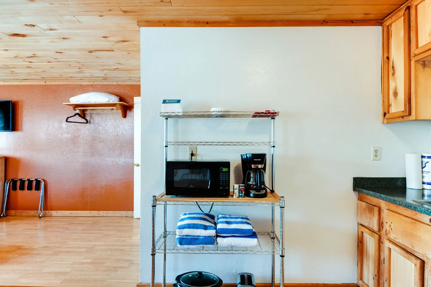 A wire shelving rack holding a microwave, coffeemaker, and towels against a white wall next to kitchen cabinets and a doorway into a living area.
