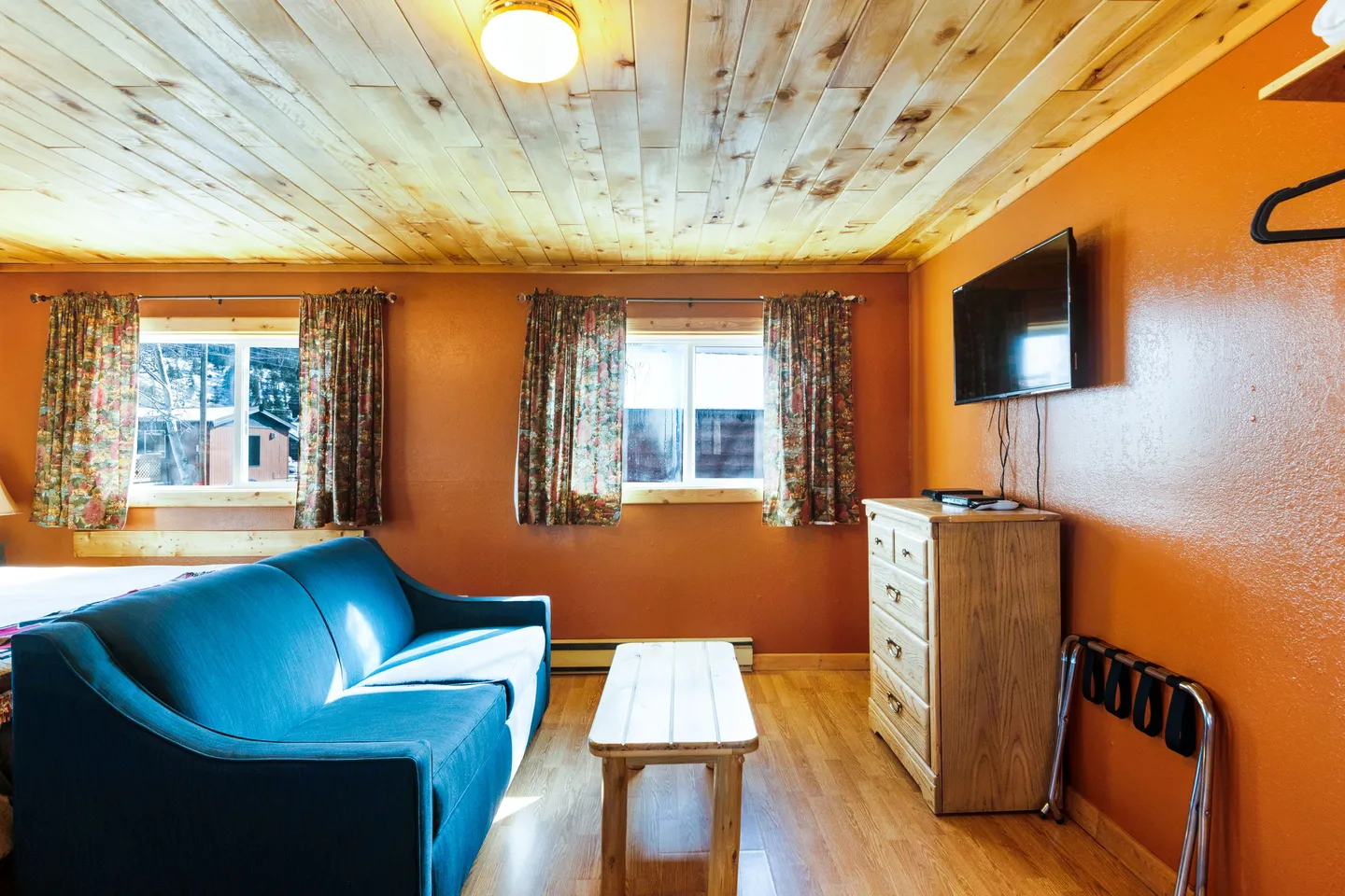 A living area with wood-plank ceiling, orange walls, blue sofa, small wooden coffee table, dresser, and wall-mounted TV.