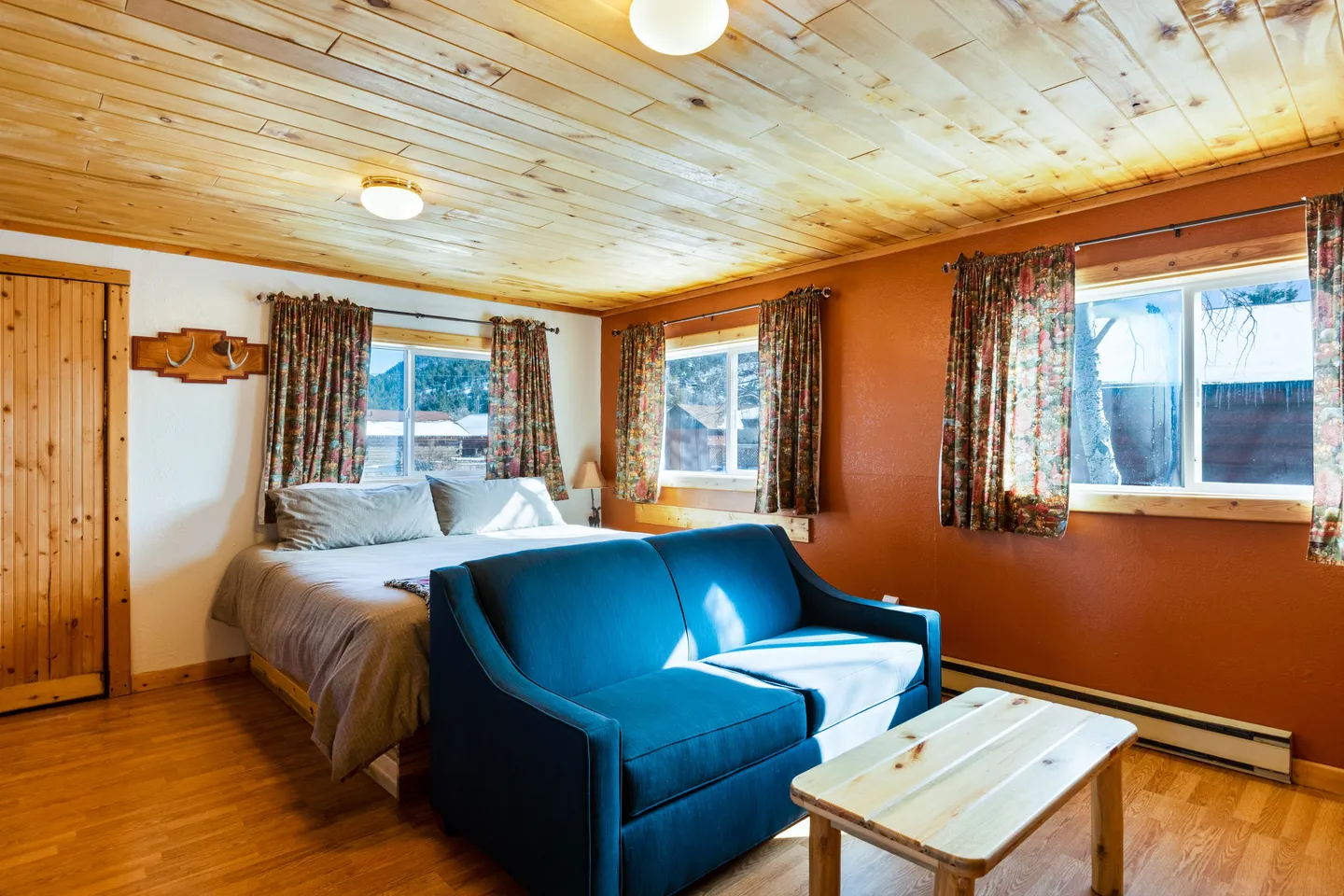Bedroom interior featuring a wood-paneled ceiling, a made bed against a windowed wall, and a blue couch with a coffee table in the foreground.