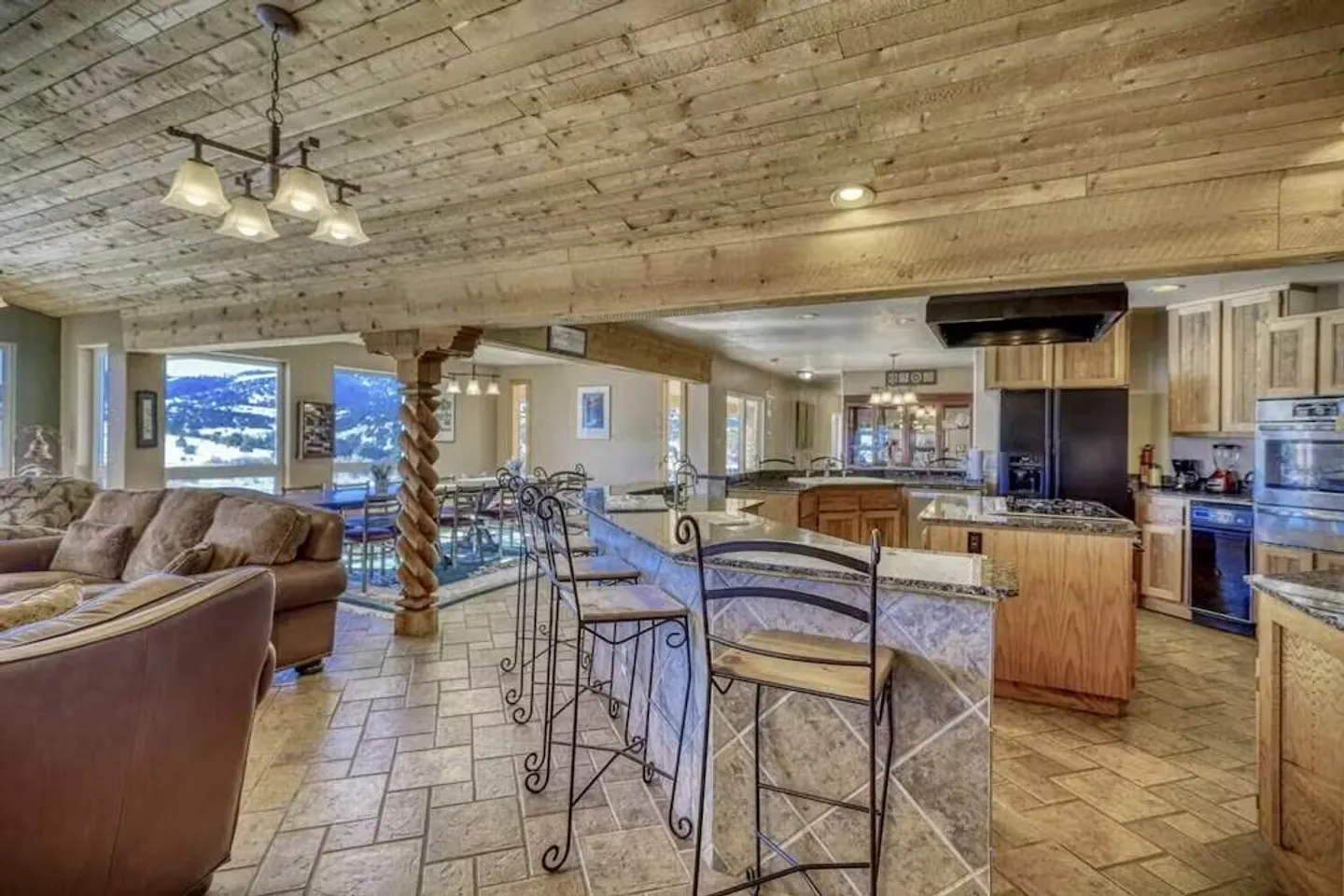 Open-concept living room and kitchen with wood-paneled ceiling, tile flooring, bar stools, and a carved wooden post.