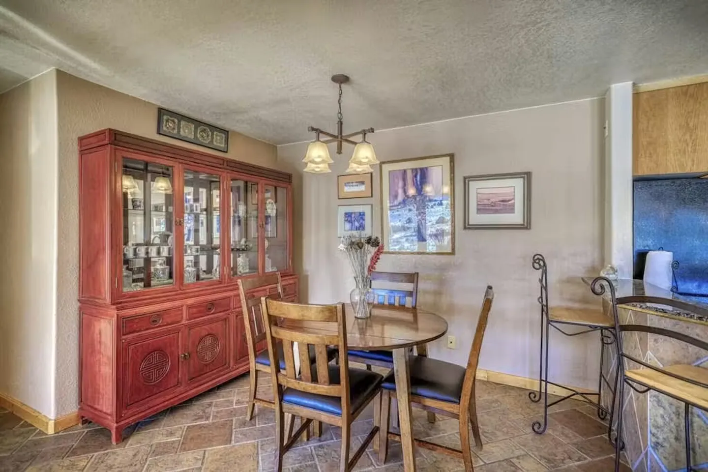 Dining area with a round wooden table, chairs, a large reddish china cabinet, and tile flooring.