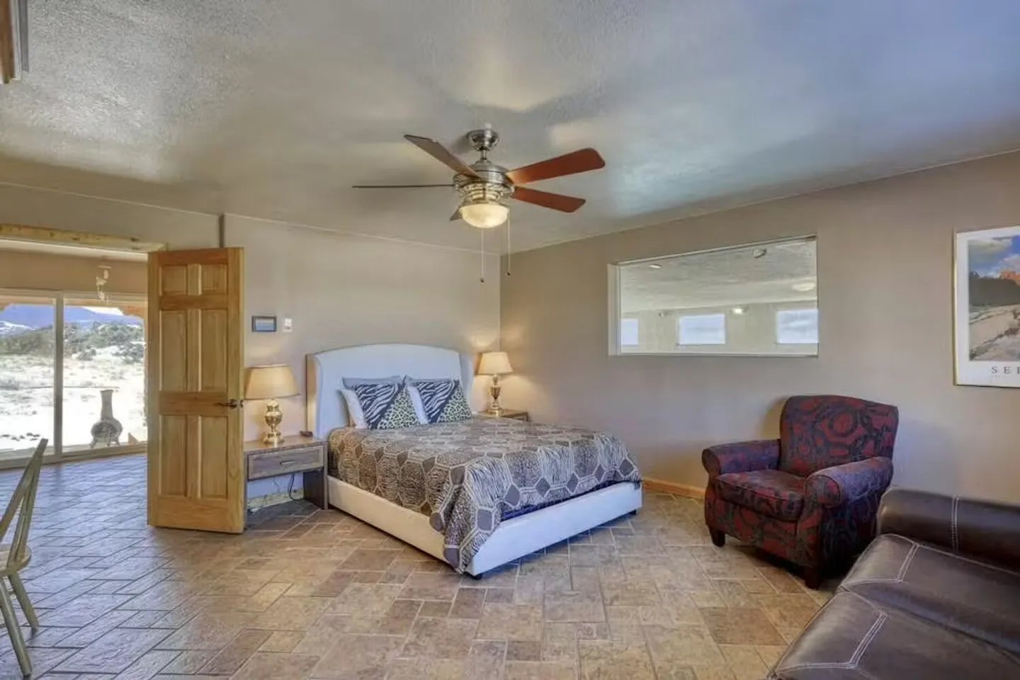 Bedroom with tile flooring, a bed with a white headboard, an armchair, ceiling fan, and sliding glass door leading outside.
