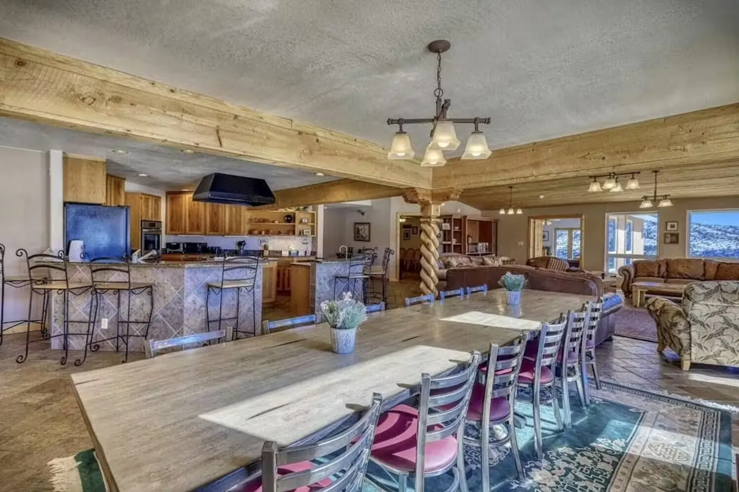 Open concept dining area, kitchen, and living room featuring exposed wood ceiling beams, a long dining table, and a kitchen island with barstools.