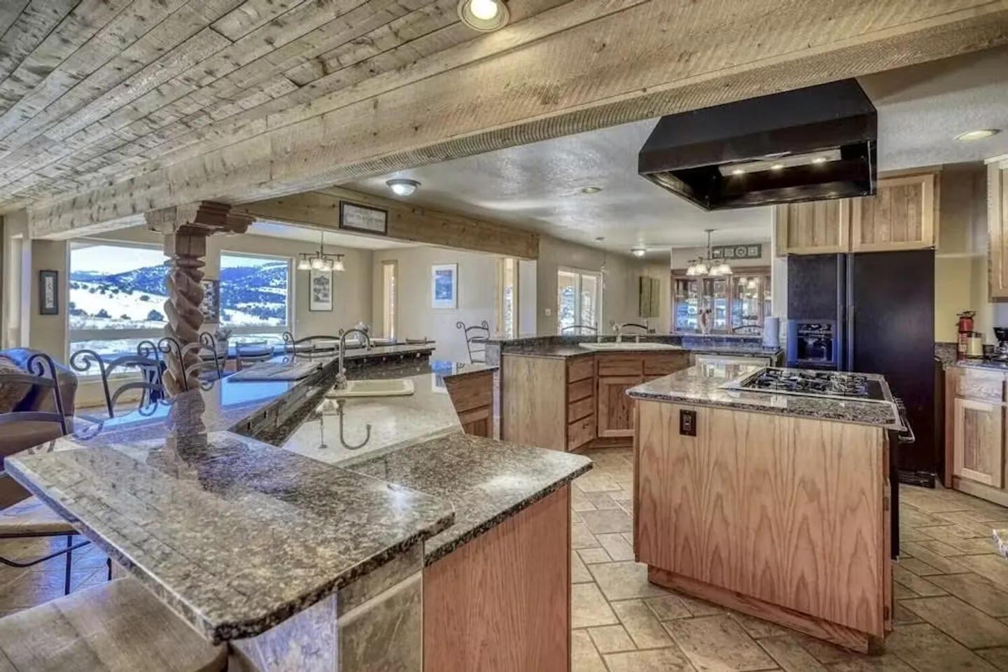 Spacious kitchen with wood cabinetry, granite-patterned countertops, an island with cooktop, and large windows framing snowy hills.