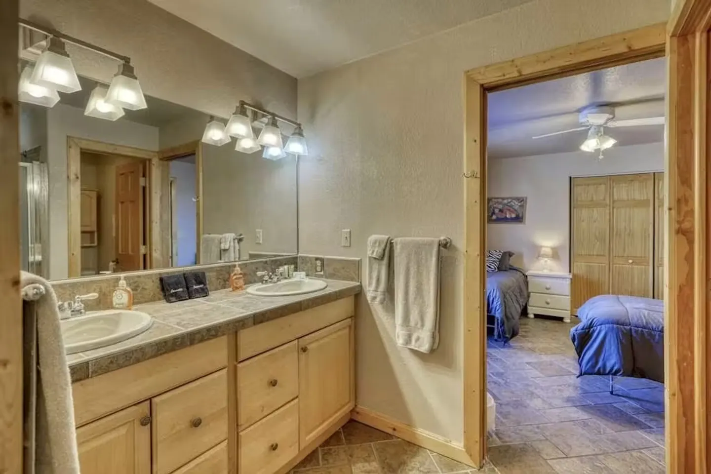 Bathroom with double vanity sinks and tiled countertop leading into an adjacent bedroom with two beds.