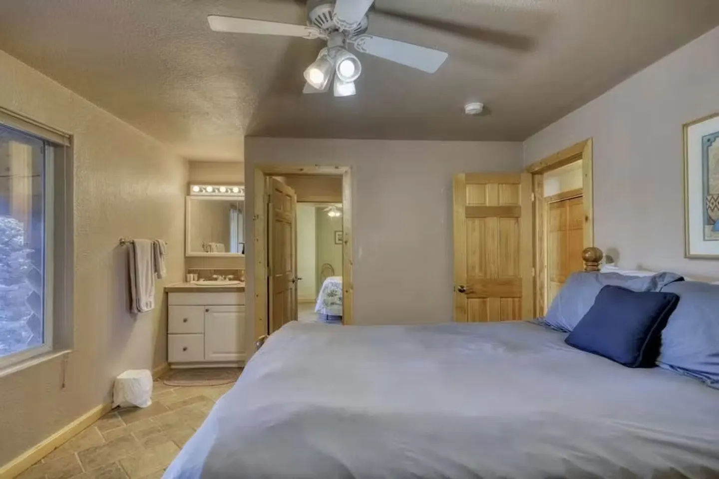 Bedroom featuring a made bed with blue bedding, a ceiling fan with lights, tiled flooring, a window, and an open bathroom vanity area.