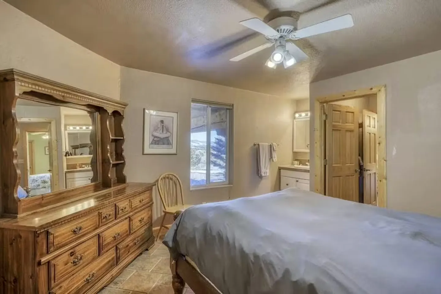 Bedroom featuring a large wooden dresser with mirror, a bed with a grey duvet, a ceiling fan, and an en-suite bathroom visible in the background.